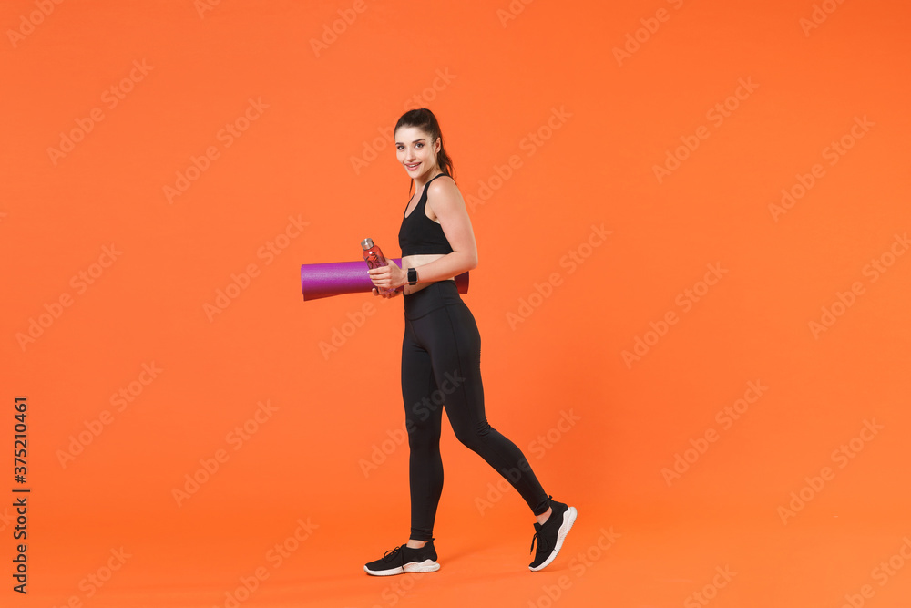 Full length side view portrait of young fitness sporty woman in black sportswear posing going training working out hold bottle of water yoga mat looking aside isolated on orange background studio.