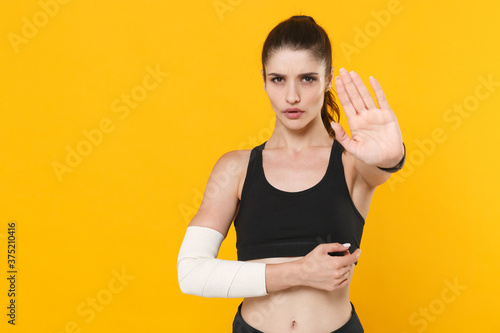 Displeased young fitness sporty woman 20s in black sportswear posing training get injured arm elbow with elastic bandage showing stop gesture with palm isolated on yellow background studio portrait.