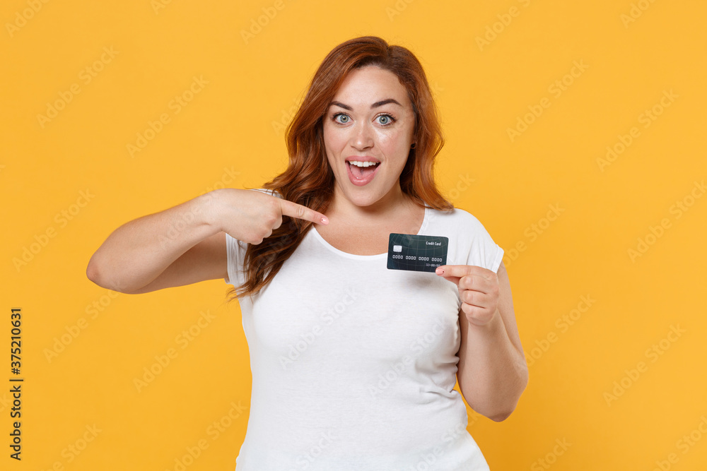 Excited young redhead plus size body positive female woman girl 20s in white blank casual t-shirt posing pointing index finger on credit bank card isolated on yellow color background studio portrait. Excited young redhead plus size body positive female woman girl 20s in white blank casual t-shirt posing pointing index finger on credit bank card isolated on yellow color background studio portrait.