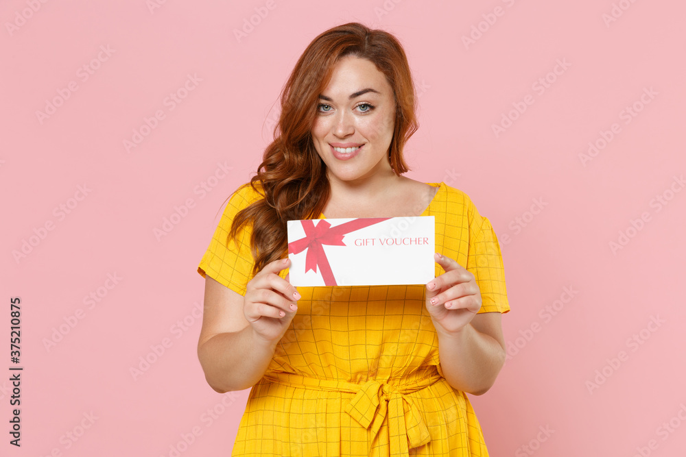 Smiling young redhead plus size body positive female woman girl 20s in yellow dress posing hold in hands gift certificate looking camera isolated on pastel pink color wall background studio portrait. Smiling young redhead plus size body positive female woman girl 20s in yellow dress posing hold in hands gift certificate looking camera isolated on pastel pink color wall background studio portrait.