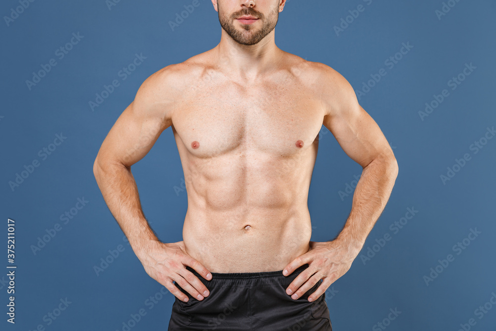 Cropped image of young bearded fitness sporty strong guy bare-chested muscular sportsman isolated on blue background. Workout sport motivation lifestyle concept. Standing with arms akimbo on waist.