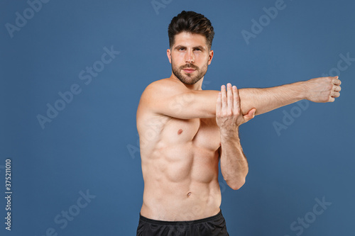 Confident young bearded fitness sporty strong guy bare-chested muscular sportsman isolated on blue background studio portrait. Workout sport motivation lifestyle concept. Doing stretching exercising.