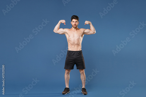Full length portrait of young bearded fitness sporty strong guy bare-chested muscular sportsman isolated on blue background studio. Workout sport motivation lifestyle concept. Showing biceps muscles.