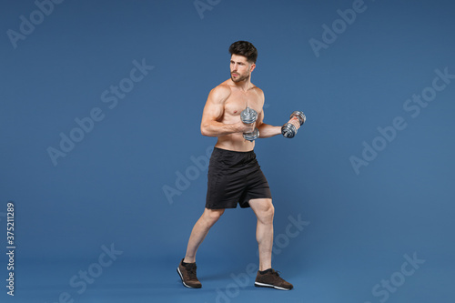 Fototapeta Naklejka Na Ścianę i Meble -  Full length portrait of young bearded fitness sporty strong guy bare-chested muscular sportsman isolated on blue background. Workout sport motivation lifestyle concept. Doing exercise with dumbbells.