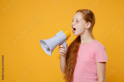 Little ginger redhead kid girl 12-13 years old wearing pink casual t-shirt posing screaming in megaphone and looking aside isolated on bright yellow color wall background children studio portrait.