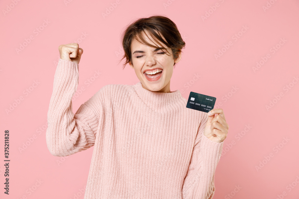 Happy young brunette woman girl wearing knitted casual sweater posing isolated on pastel pink wall background studio portrait. People lifestyle concept. Hold credit bank card, doing winner gesture. Happy young brunette woman girl wearing knitted casual sweater posing isolated on pastel pink wall background studio portrait. People lifestyle concept. Hold credit bank card, doing winner gesture.