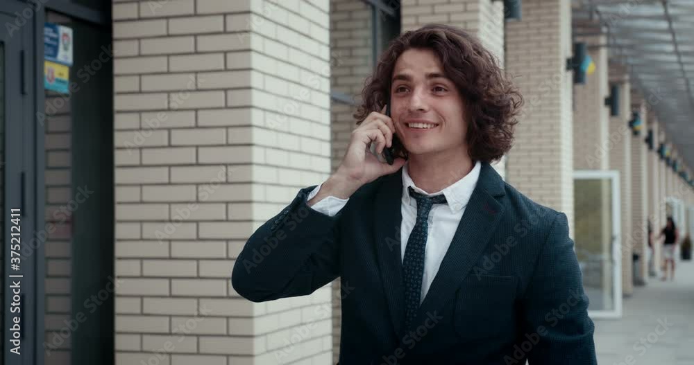 Successful young businessman walking outdoors talking on phone, having pleasant conversation, looking satisfied and in good mood. Successful young businessman walking outdoors talking on phone, having pleasant conversation, looking satisfied and in good mood.