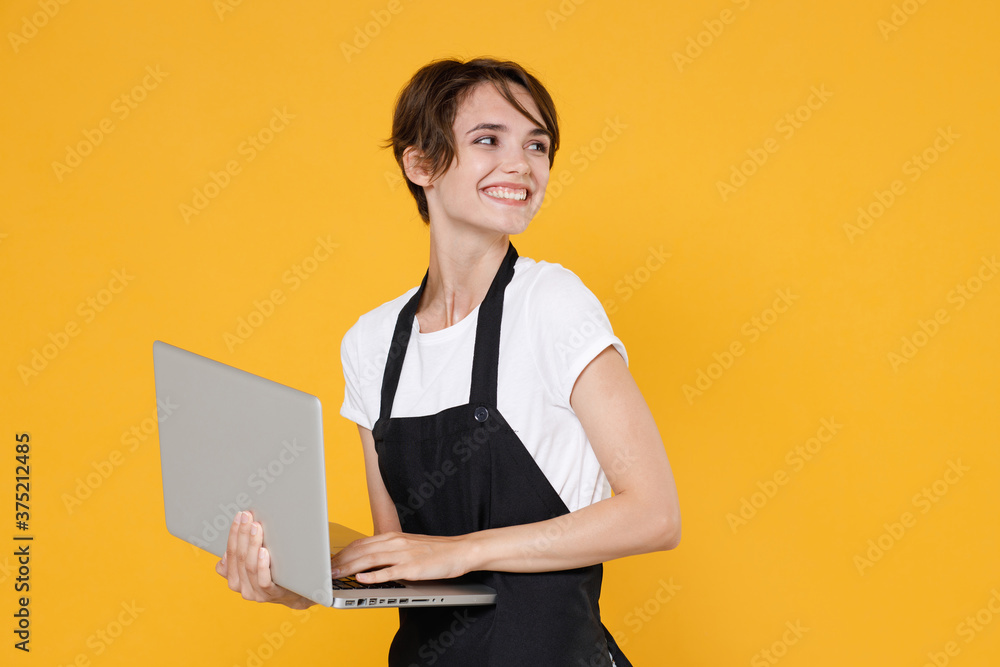 Smiling young female woman 20s barista bartender barman employee in white t-shirt apron posing working on laptop pc computer looking aside isolated on yellow color wall background studio portrait. Smiling young female woman 20s barista bartender barman employee in white t-shirt apron posing working on laptop pc computer looking aside isolated on yellow color wall background studio portrait.