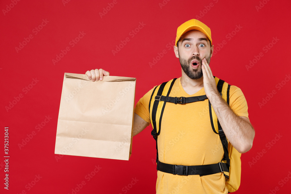 Delivery man in yellow cap t-shirt thermal backpack hold brown clear empty blank craft paper food takeaway bag mock up isolated on red background studio Guy male employee work courier Service concept