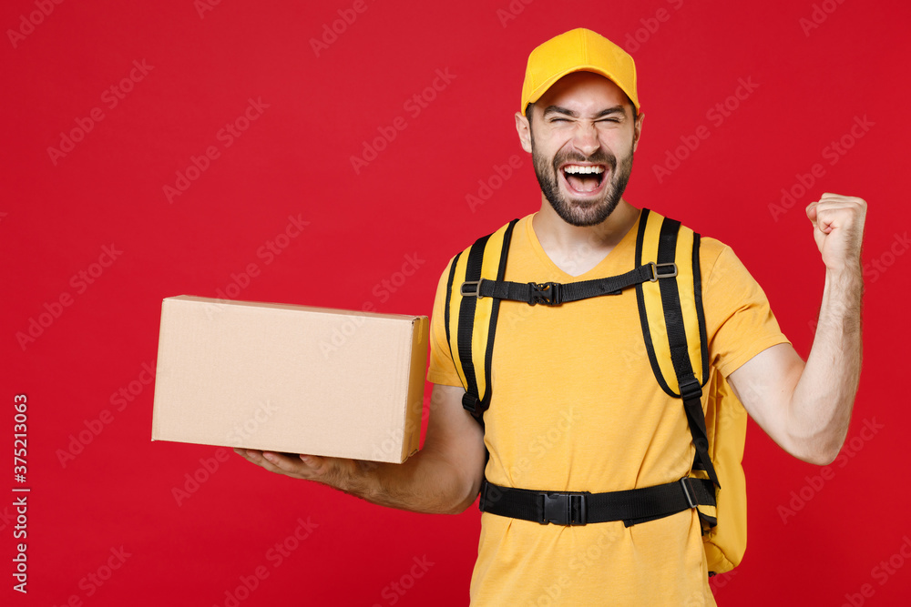 Delivery man in yellow cap t-shirt uniform thermal bag backpack with food hold cardboard box isolated on red background studio Male employee work as courier Service coronavirus covid-19 virus concept