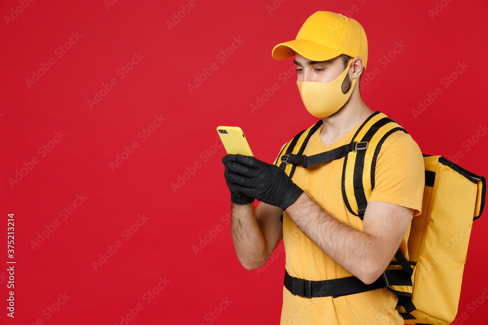 Delivery employee man in yellow cap face mask gloves t-shirt uniform thermal food bag backpack, hold cellphone, work courier service during quarantine covid-19 virus isolated on red background studio.