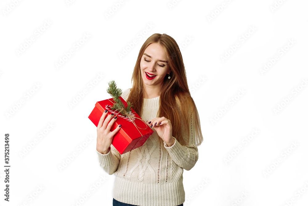 Surprised young woman holds in a white sweater holding a Christmas red gift with a fir branch and a festive bow. The happy model is happy to receive a gift and opens her mouth in surprise Surprised young woman holds in a white sweater holding a Christmas red gift with a fir branch and a festive bow. The happy model is happy to receive a gift and opens her mouth in surprise