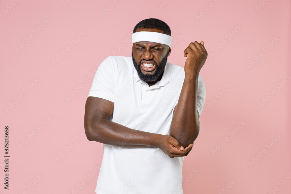 Sick injured young bearded african american fitness sports man in headband t-shirt feels bad pain spasm touching elbow arm spending time in gym isolated on pink color wall background studio portrait. Sick injured young bearded african american fitness sports man in headband t-shirt feels bad pain spasm touching elbow arm spending time in gym isolated on pink color wall background studio portrait.