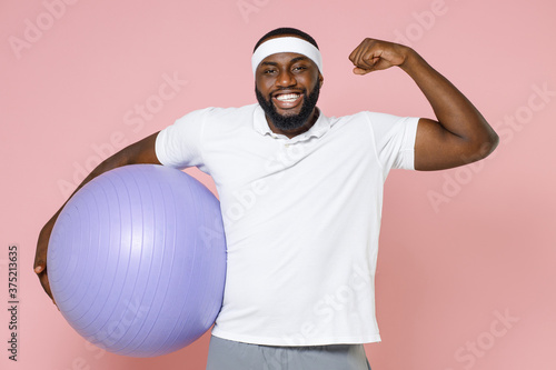 Smiling young bearded african american fitness sports man in white headband t-shirt hold fitball showing biceps muscles spending time in gym isolated on pastel pink color background studio portrait.