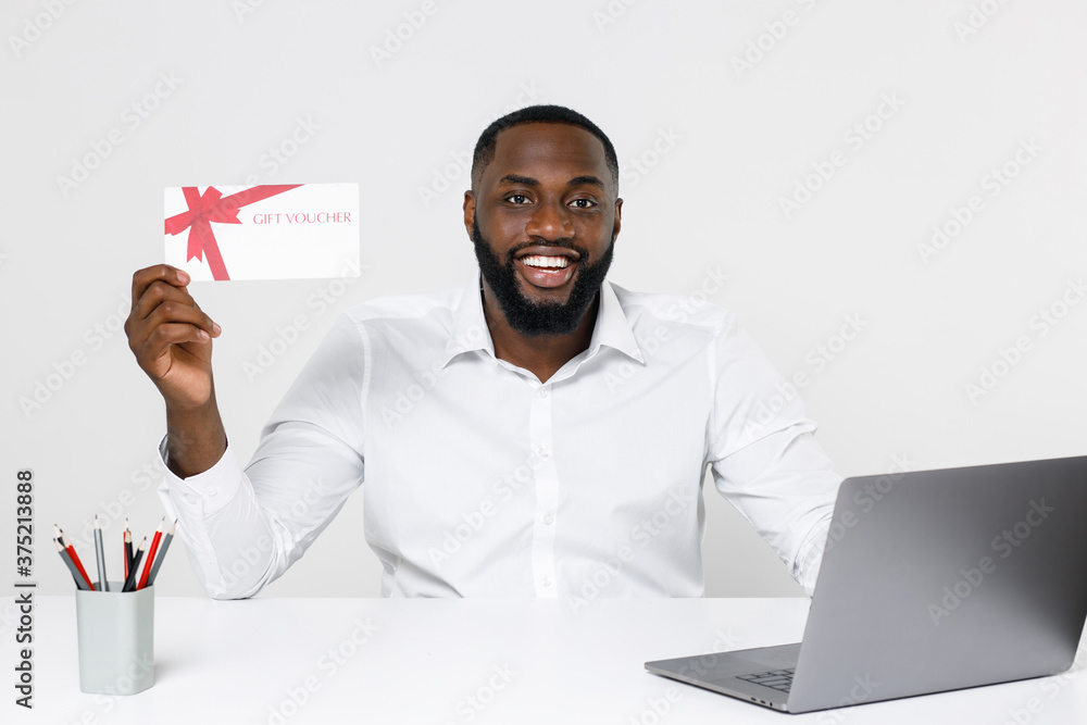 Smiling young african american male business man in classic white shirt posing working in office sitting at desk with laptop pc hold gift certificate looking camera isolated on white color background. Smiling young african american male business man in classic white shirt posing working in office sitting at desk with laptop pc hold gift certificate looking camera isolated on white color background.