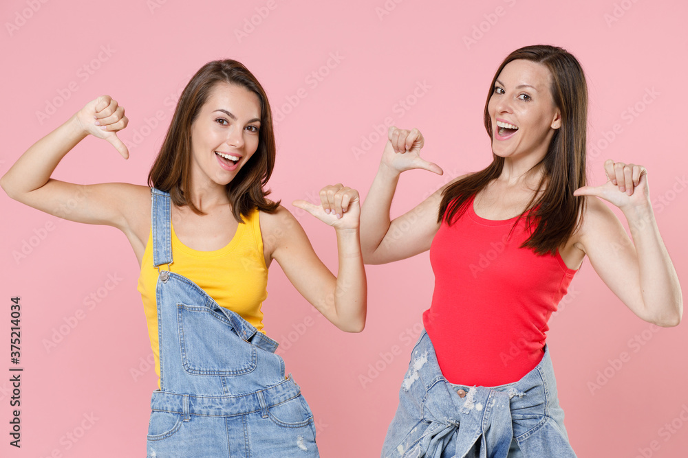 Two excited cheerful funny attractive young women friends 20s wearing casual denim clothes pointing thumbs on themselves looking camera isolated on pastel pink colour background, studio portrait. Two excited cheerful funny attractive young women friends 20s wearing casual denim clothes pointing thumbs on themselves looking camera isolated on pastel pink colour background, studio portrait.