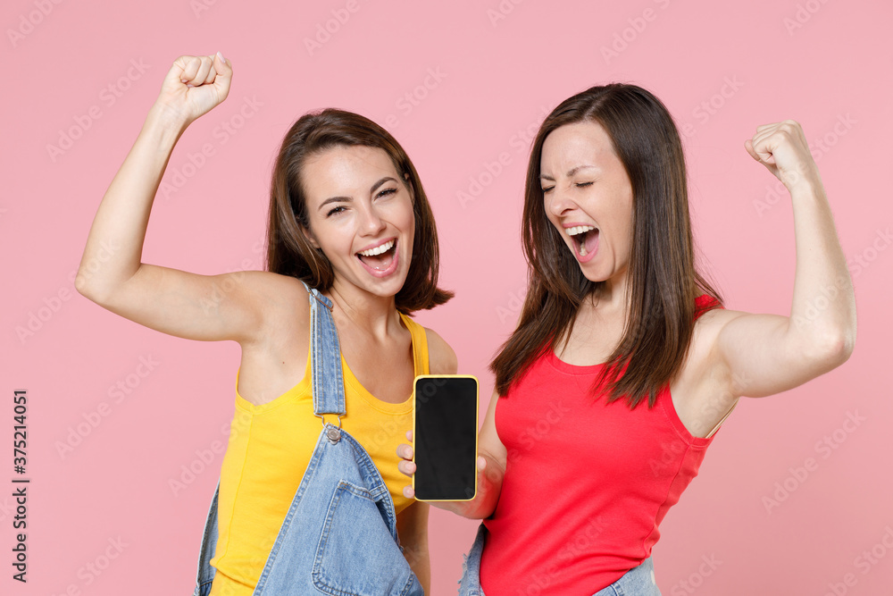 Two happy joyful young women friends in denim clothes doing winner gesture hold mobile phone with blank empty screen mock up copy space isolated on pastel pink colour background, studio portrait. Two happy joyful young women friends in denim clothes doing winner gesture hold mobile phone with blank empty screen mock up copy space isolated on pastel pink colour background, studio portrait.