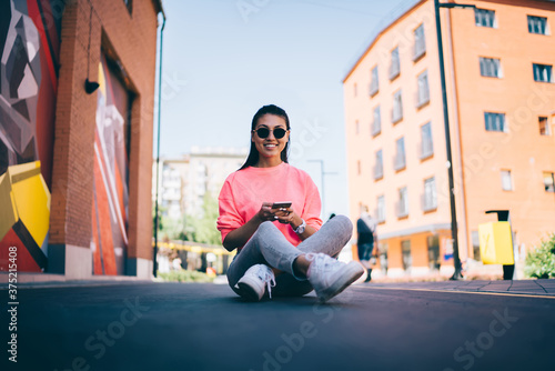 Toothy hipster girl in stylish sunglasses enjoying leisure for networking smartphone browser, prosperous Asian millennial 20s with cellphone technology resting in city sitting at asphalt road