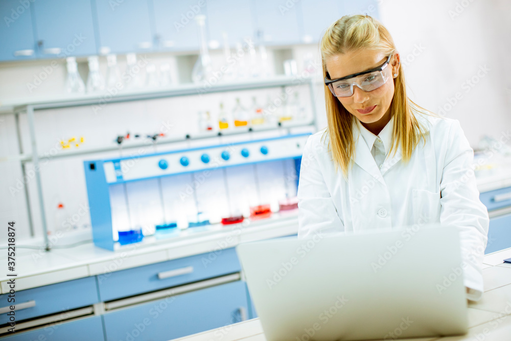 Female researcher in white lab coat and protective goggles using laptop while working in the laboratory Female researcher in white lab coat and protective goggles using laptop while working in the laboratory