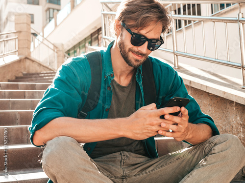 Handsome stylish smiling hipster lambersexual model.Man dressed in white T-shirt.Fashion male sitting on the stairs. Holding, scrolling and texting in his cellphone. Writes sms on smartphone photo