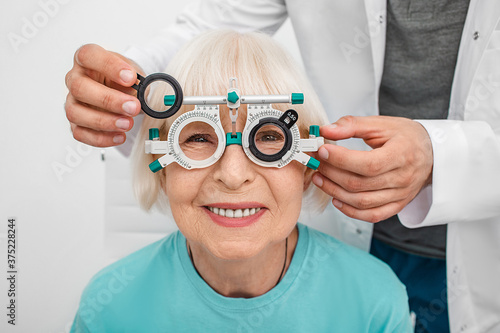 Smiling senior woman wearing optometrist trial frame at ophthalmology clinic. Ophthalmologist helping select glasses for treatment of vision. Eye check-up