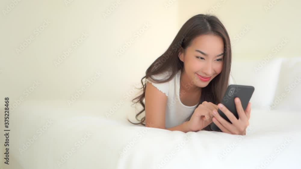 Simplistic white concept, woman typing on her mobile phone while lying on the cozy king-size bed facing the camera smiling, camera slide up Simplistic white concept, woman typing on her mobile phone while lying on the cozy king-size bed facing the camera smiling, camera slide up