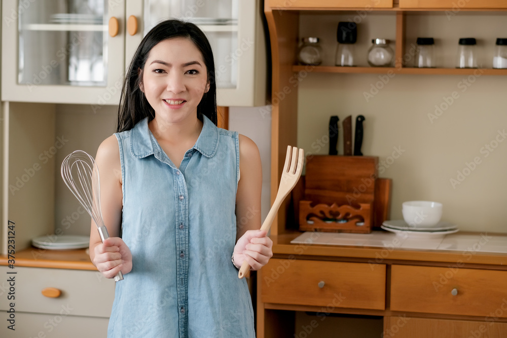 Asian woman hold whisk and fork stand in kitchen with day light and she look happy. Concept of preparation for cooking in her home. Asian woman hold whisk and fork stand in kitchen with day light and she look happy. Concept of preparation for cooking in her home.