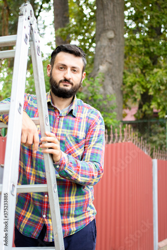 Young brunette bearded man standing on ladder on background of fence and trees in the backyard, wearing red green checkered shirt and looking at camera. Repair, yard work, gardening concept.Vertical