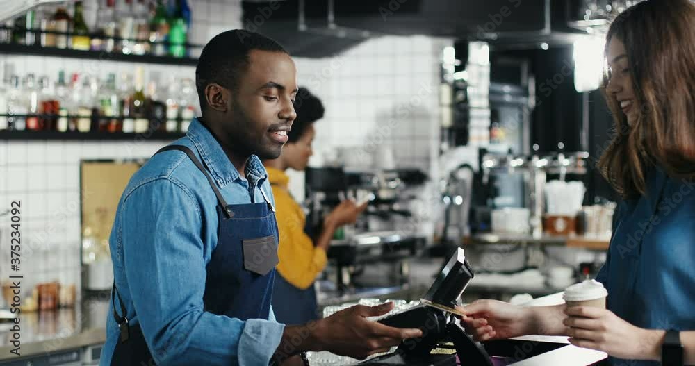 Multiethnic bartender man and client woman. Caucasian female paying with paypass, credit card. Mixed-races. Passing payment at counter. Pay for drink of coffee. African American waiter. Multiethnic bartender man and client woman. Caucasian female paying with paypass, credit card. Mixed-races. Passing payment at counter. Pay for drink of coffee. African American waiter.