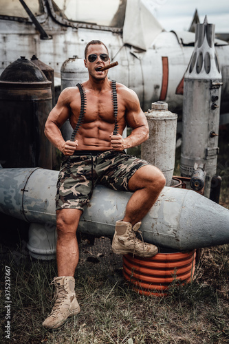 Cool macho with camouflage shorts relaxing and smoking cigar. Abandoned old airplane in the field and brutal military with sunglasses.