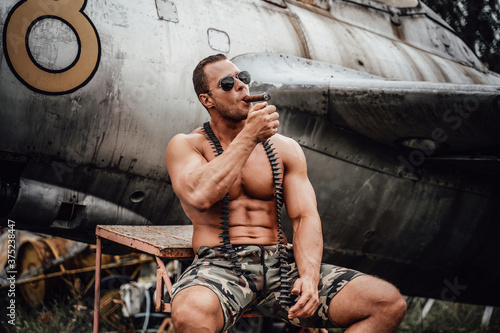 Old war airplane in the abandoned airport and brutal naked soldier near it. American bodybuilder smoking cuban cigar in the field.