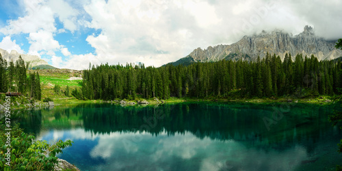 Panoramic view of Lago di Carezza, pine forest and Catinaccio Mountains reflected on the clear water on a cloudy summer day, Sudtirol, Trentino Alto Adige, Dolomites, Unesco, Italy