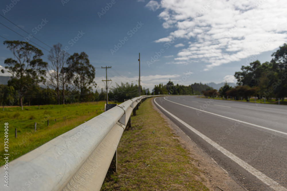 Closeup to a metal safety highway bar in a country field highway curve with green trees and blue cloudy sky in the morning 