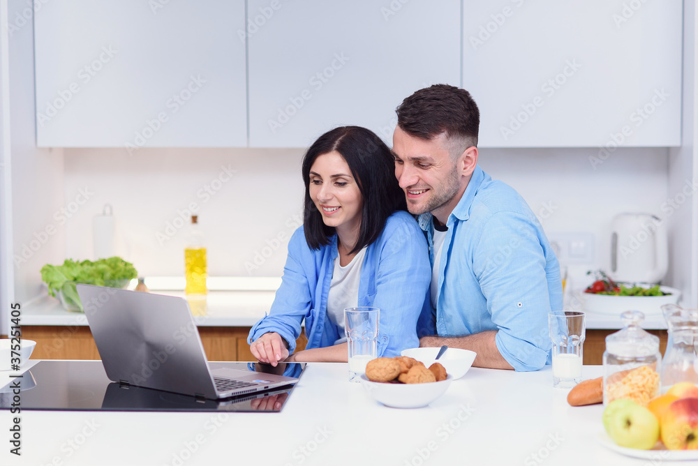Beautiful happy young couple having breakfast at home and uses laptop at kitchen. Enjoying free time together. Beautiful happy young couple having breakfast at home and uses laptop at kitchen. Enjoying free time together.