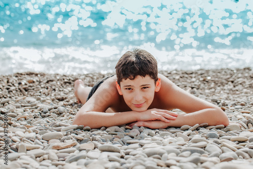 Beautiful brunette boy sunbathes and smiles lying on pebble beach against the sea