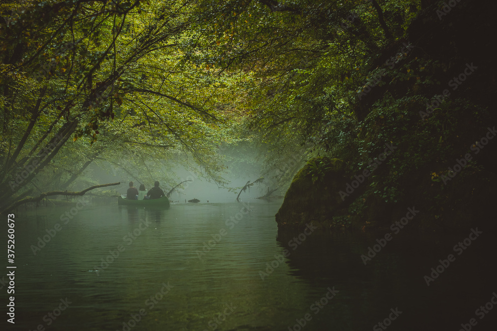 Fototapeta premium Back view of a green canoe and people in a misty and foggy river between the trees. Scary spooky and mistery exploration with a canoe on a river Krupa in Slovenia.