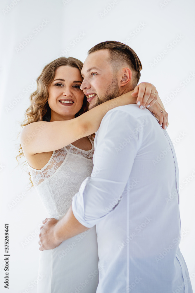 Fototapeta premium A beautiful curly pregnant girl in an evening dress stands in an embrace with her husband on a white background. She has a white evening dress and he is wearing nice white shirt. (selective focus)