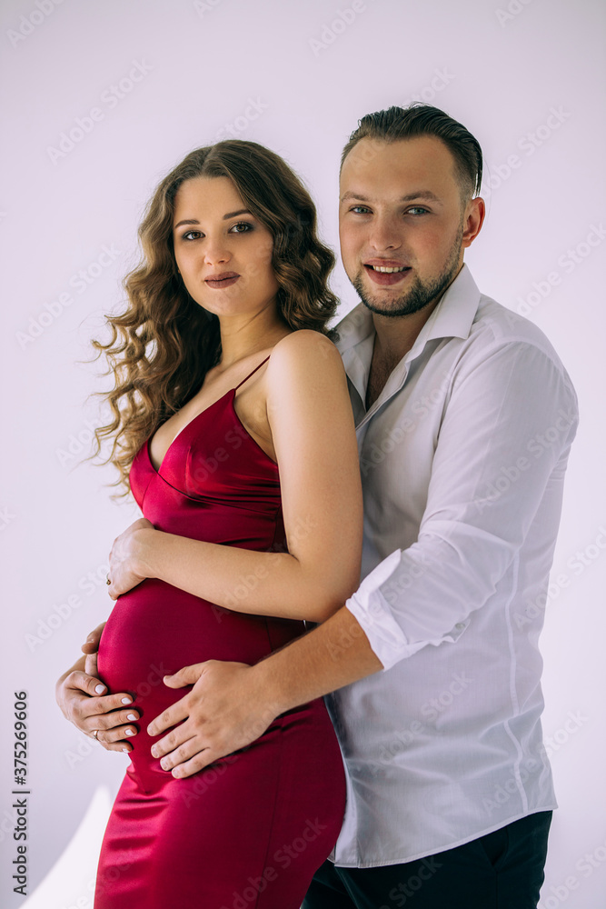 A beautiful curly pregnant girl in an evening dress stands in an embrace with her husband on a white background. She has a crimson evening dress and he is wearing festive white shirt. Family Concept A beautiful curly pregnant girl in an evening dress stands in an embrace with her husband on a white background. She has a crimson evening dress and he is wearing festive white shirt. Family Concept