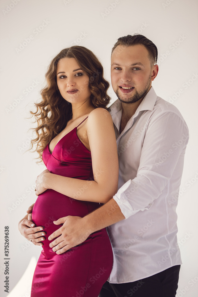 A beautiful curly pregnant girl in an evening dress stands in an embrace with her husband on a white background. She has a crimson evening dress and he is wearing festive white shirt. Family Concept A beautiful curly pregnant girl in an evening dress stands in an embrace with her husband on a white background. She has a crimson evening dress and he is wearing festive white shirt. Family Concept
