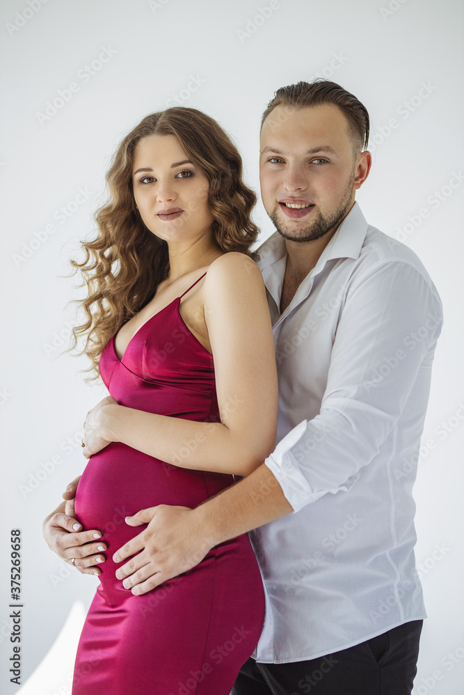 A beautiful curly pregnant girl in an evening dress stands in an embrace with her husband on a white background. She has a crimson evening dress and he is wearing festive white shirt. Family Concept A beautiful curly pregnant girl in an evening dress stands in an embrace with her husband on a white background. She has a crimson evening dress and he is wearing festive white shirt. Family Concept