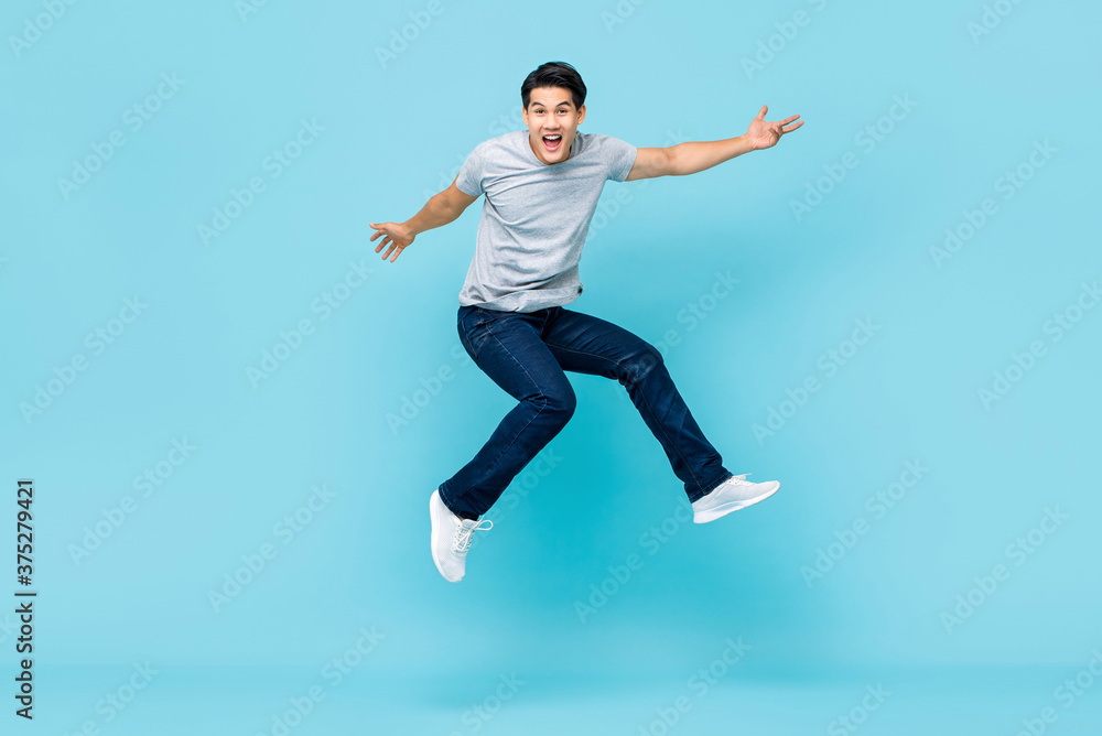 Energetic happy young Asian man in casual clothes jumping studio shot isolated in light blue background