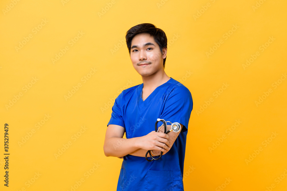 Young handsome Asian male doctor smiling and doing arms crossed gesture isolated on yellow background Young handsome Asian male doctor smiling and doing arms crossed gesture isolated on yellow background