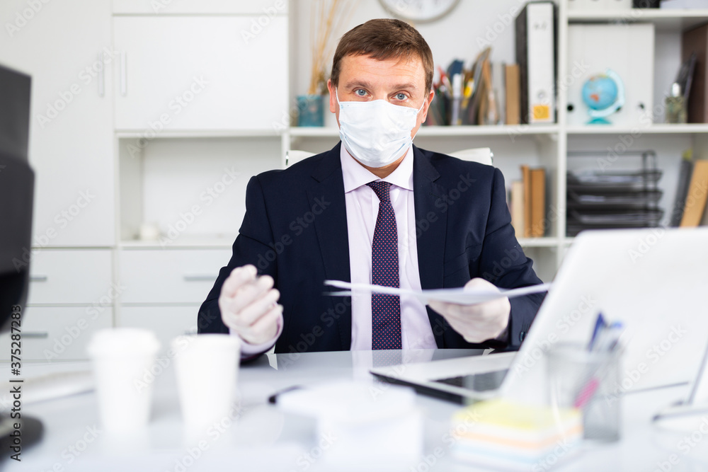 Male worker wearing medical facial mask and gloves engaged in business activities at table in office