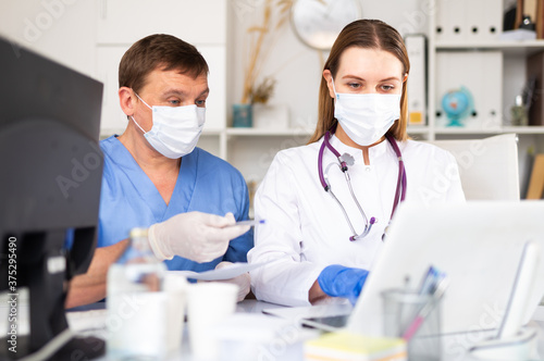 Experienced adult doctor helping young female colleague to fill up medical forms in laptop