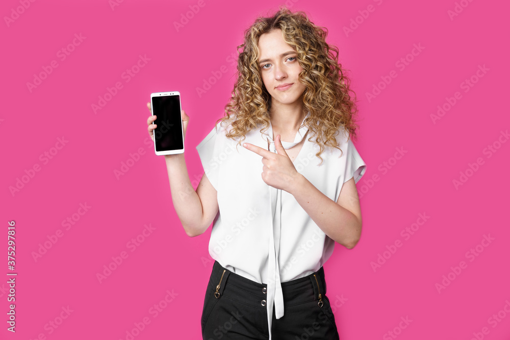Look at this cell phone. Contented happy woman, pointing her index finger at a blank screen, shows a modern device. Isolated on a pink background. Look at this cell phone. Contented happy woman, pointing her index finger at a blank screen, shows a modern device. Isolated on a pink background.