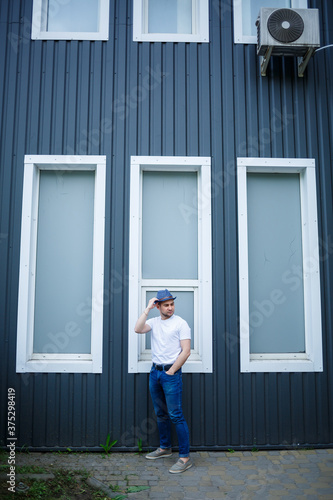 A cool young man in a stylish white t-shirt and hat stands near a gray wall with large windows in the city. Attractive modern guy. Fashionable summer men's clothing. Street style © Дмитрий Ткачук