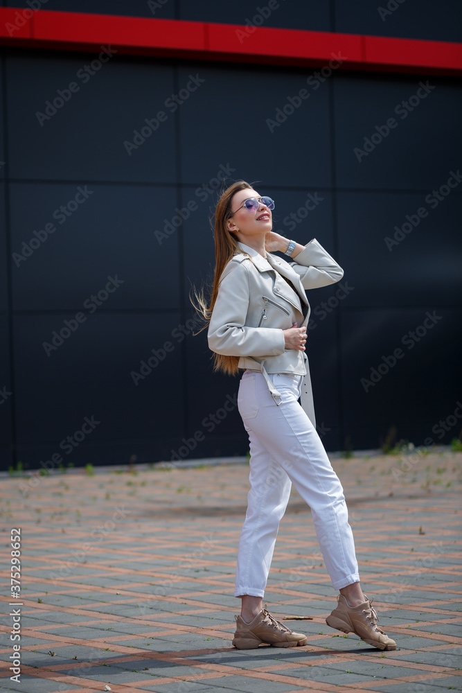 Photo of a long-haired girl in a white blouse and light jeans stands with a smile on the background of the gray wall of the building on a sunny spring day.