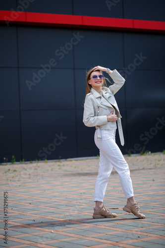 Photo of a long-haired girl in a white blouse and light jeans stands with a smile on the background of the gray wall of the building on a sunny spring day.