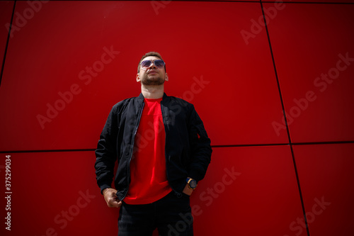 A cool young man in trendy sunglasses in a stylish red T-shirt with a black jacket stands near a red building in the city. Attractive modern guy. Fashionable summer men's clothing. Street style