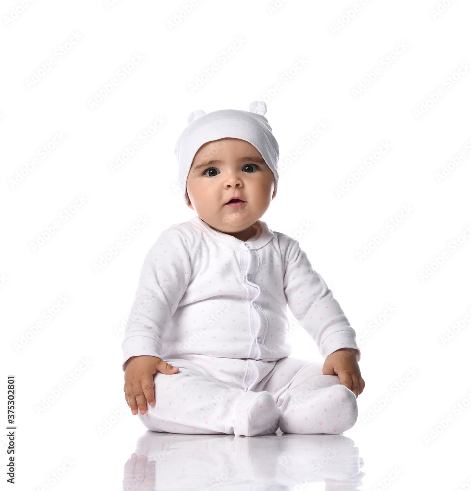 Cute infant baby toddler sits on the floor in white jumpsuit overall and funny hat with ears holding hands at knees Cute infant baby toddler sits on the floor in white jumpsuit overall and funny hat with ears holding hands at knees